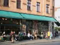 Menschen sitzen an Tischen vor einem Café in Berlin. Die Fassade ist mit Graffiti verziert und trägt die Aufschrift "dorle berlin".