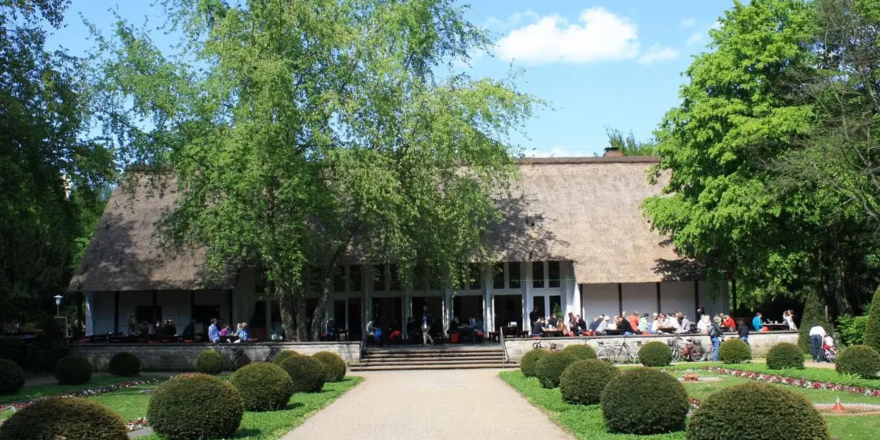 Ein idyllischer Tag im Englischen Garten Berlin. Menschen genießen das Wetter vor einem Gebäude mit Reetdach, umgeben von grünen Bäumen und gepflegten Büschen.
