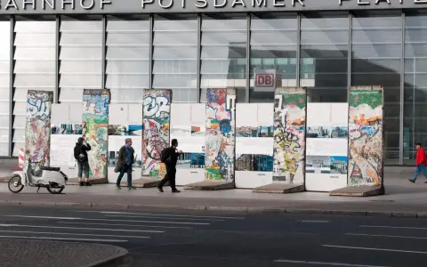 Bunte Reste des Berliner Mauer-Denkmals vor dem Bahnhof Potsdamer Platz. Menschen spazieren vorbei, ein Roller parkt daneben.