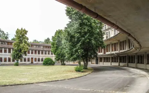 Alte Gebäude im olympisches Dorf Berlin, umgeben von Bäumen und Grünflächen. Eine Straße führt an einem geschwungenen Gebäude mit vielen Fenstern vorbei.