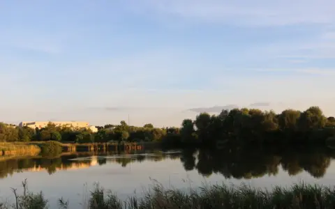 Spiegelglatte Wasserfläche am Wuhletal Wanderweg, reflektiert Bäume und Gebäude. Ein rotes Haus sticht hervor.
