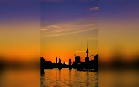 Silhouetten der Berliner Skyline spiegeln sich im Wasser. Ein dramatischer Sonnenuntergang taucht den Himmel in Orange- und Violetttöne.