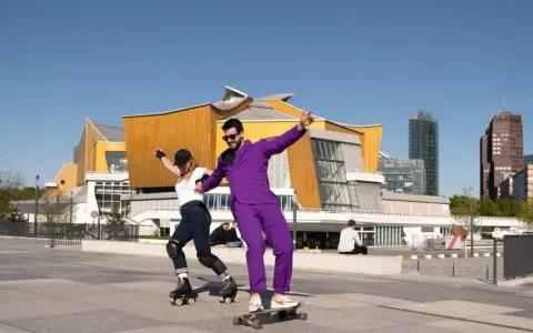 Ein Mann im lila Anzug auf einem Skateboard und eine Frau auf Rollschuhen tanzen vor der Philharmonie. Ein sonniger Tag heute in Berlin.