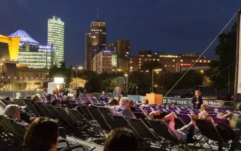 Leute entspannen sich in Liegestühlen beim **Sommerkino am Potsdamer Platz** vor einer leuchtenden Skyline.