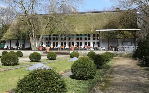 Das Teehaus im Englischen Garten Berlin mit seinem Reetdach und der Terrasse, auf der Gäste sitzen, ist von gepflegten Rasenflächen und Bäumen umgeben.