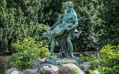 Bronzestatue eines Mannes, der eine Frau trägt, vor einem grünen Hintergrund mit einem kleinen Wasserfall in Berlin.