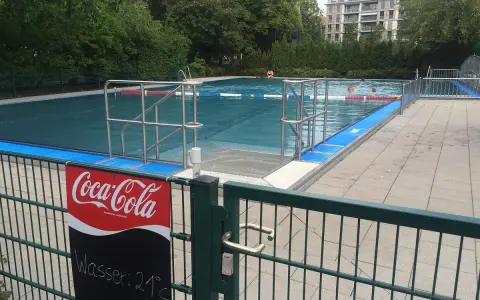 Erfrischung im Monbijou Bad: Wasser 21°C. Menschen schwimmen im blauen Becken, umgeben von Bäumen und einem modernen Gebäude.