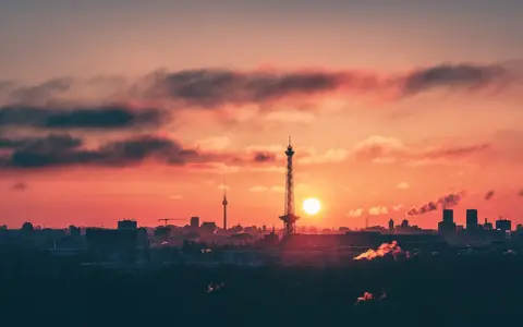 Berlin bei Nacht von oben: Die Sonne geht unter und taucht den Himmel in ein feuriges Rot. Silhouetten von Türmen und Gebäuden zeichnen sich ab.
