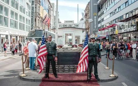 Zwei Männer in Uniformen mit US-Flaggen stehen am Checkpoint Charlie in Berlin. Touristen und ein McDonald's im Hintergrund.