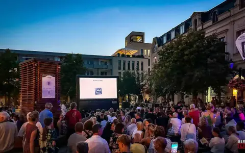 Ein lauer Sommerabend in Berlin: Menschen genießen ein Open-Air-Kino-Erlebnis vor einer Leinwand mit dem Logo "Lux Augenoptik".