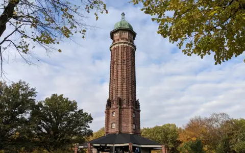 Der Aussichtsturm im Volkspark Jungfernheide ragt mit seinem roten Backstein und grünen Kuppeldach in den Himmel. Herbstlaub umrahmt die Szene.