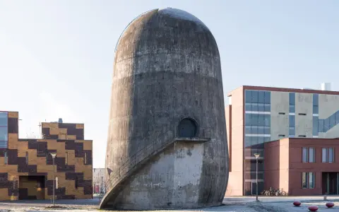 Der Trudelturm Berlin, ein futuristisches Betonbauwerk, steht neben modernen Gebäuden. Eine Außentreppe führt zu einer runden Tür.