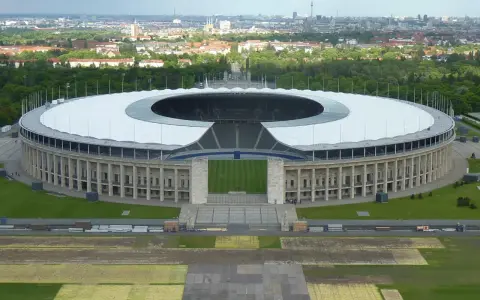 Das Olympiastadion Berlin mit seinem markanten Dach und der Skyline im Hintergrund. In der Ferne ragt der Fernsehturm, der wie ein glockenturm Berlin überragt.