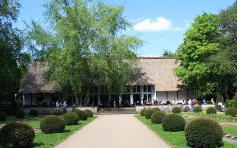 Ein idyllischer Tag im Englischen Garten Berlin. Menschen genießen das Wetter vor einem Gebäude mit Reetdach, umgeben von grünen Bäumen und gepflegten Büschen.