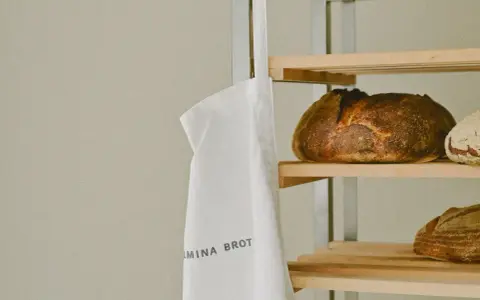 Ein weißer Stoffbeutel mit der Aufschrift "WILMINA BROT" hängt neben einem Regal mit frisch gebackenen Broten.