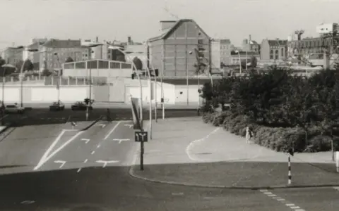 Straßenszene in Berlin der 80er Jahre mit Autos, Ampeln und einem Kran im Hintergrund.