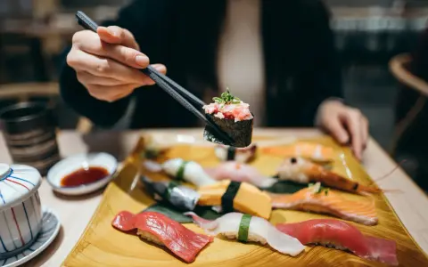 Eine Hand hält mit Stäbchen ein Stück Sushi mit gehacktem Thunfisch. Auf dem Teller liegen verschiedene Sushi-Rollen und Nigiri, bereit für den Genuss in Berlin.
