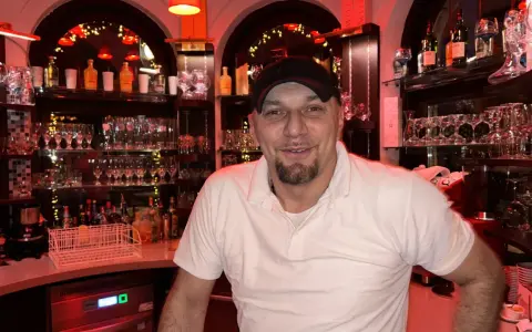 Mann mit Bart und Kappe hinter der Bar, umgeben von Gläsern und Flaschen. Er lächelt freundlich, bereit für die nächste Bestellung in "la gondola due".