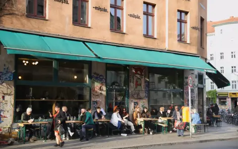 Menschen sitzen an Tischen vor einem Café in Berlin. Die Fassade ist mit Graffiti verziert und trägt die Aufschrift "dorle berlin".