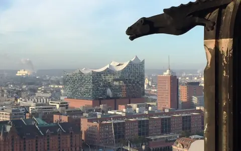 Blick über Hamburg mit der Elbphilharmonie, gesehen durch ein Fenster des Mahnmals St. Nikolai. Ein Wasserspeier wacht über die Stadt.