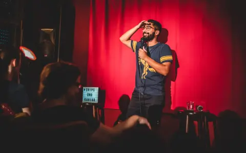 Ein Comedian steht auf einer Bühne in Berlin, hält ein Mikrofon und wischt sich die Stirn. Ein Schild mit "Chips und Kaviar" steht daneben.