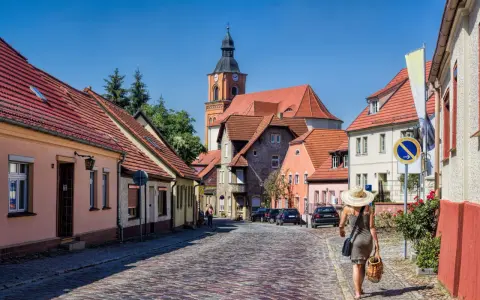 Frau mit Strohhut schlendert durch eine malerische Gasse mit Kopfsteinpflaster und roten Ziegeldächern. Ein besonderes Flair in Berlin.