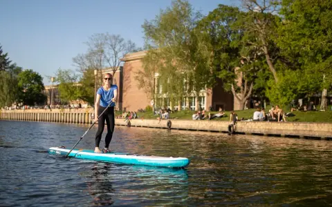 Frau genießt Stand Up Paddling auf einem Fluss, während Menschen am Ufer entspannen.