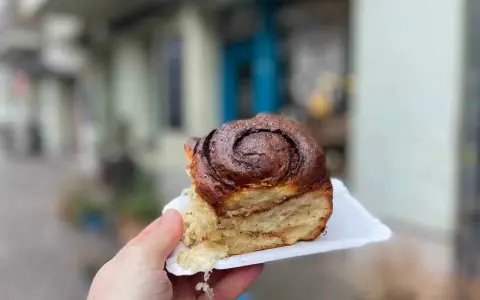 Hand hält ein saftiges Zimtschnecken-Stück vor einer unscharfen Berliner Kulisse.