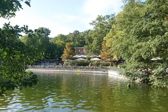 Grüne Bäume säumen den Schlachtensee Berlin, wo Menschen auf der Terrasse eines Restaurants sitzen.