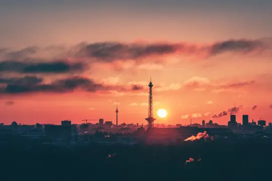 Berlin bei Nacht von oben: Die Sonne geht unter und taucht den Himmel in ein feuriges Rot. Silhouetten von Türmen und Gebäuden zeichnen sich ab.