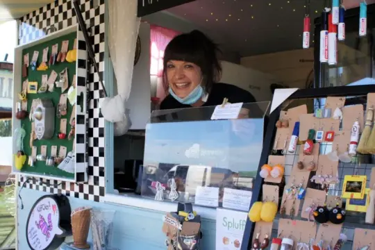 Lachende Verkäuferin auf dem Flohmarkt Friedrichshagen präsentiert handgefertigten Schmuck, darunter Eulen-Ohrringe und Miniatur-Flaschen.