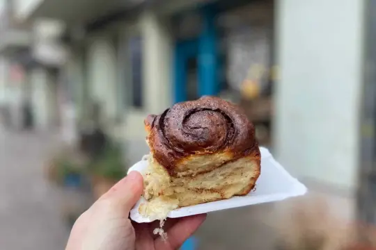 Hand hält ein saftiges Zimtschnecken-Stück vor einer unscharfen Berliner Kulisse.