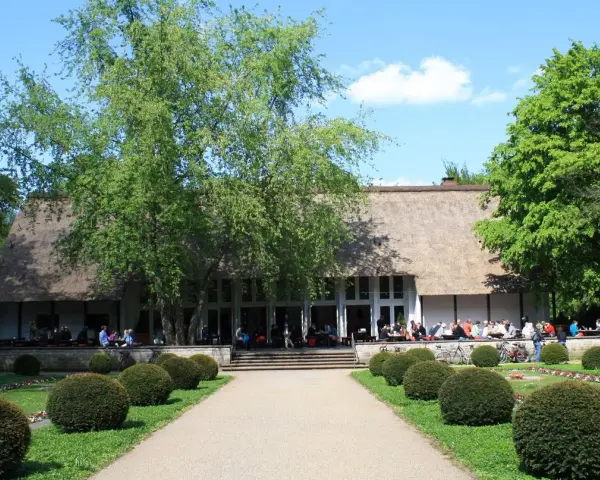 Ein idyllischer Tag im Englischen Garten Berlin. Menschen genießen das Wetter vor einem Gebäude mit Reetdach, umgeben von grünen Bäumen und gepflegten Büschen.