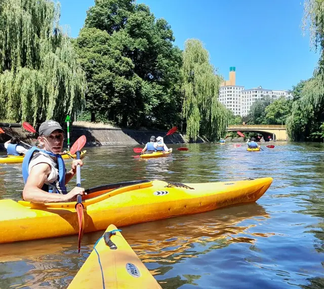 Eine Gruppe von Menschen paddelt auf gelben Kajaks auf einem Fluss in Berlin. Bäume säumen das Ufer, im Hintergrund ist ein Gebäude zu sehen.