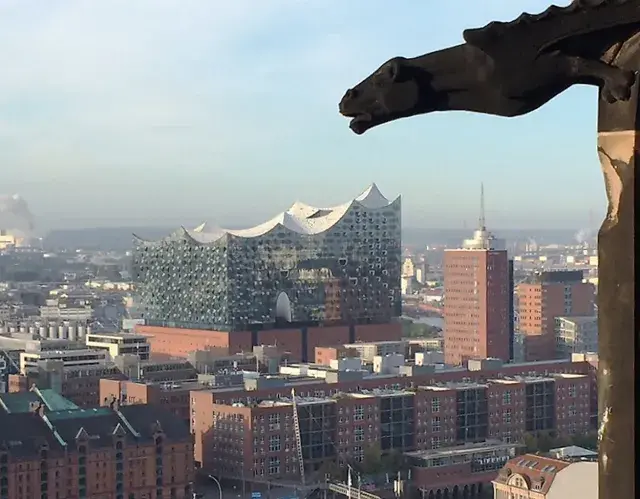 Blick über Hamburg mit der Elbphilharmonie, gesehen durch ein Fenster des Mahnmals St. Nikolai. Ein Wasserspeier wacht über die Stadt.