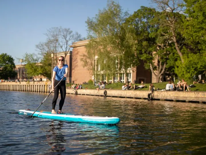 Frau genießt Stand Up Paddling auf einem Fluss, während Menschen am Ufer entspannen.