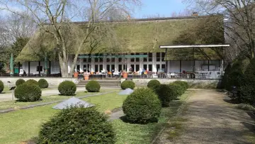 Das Teehaus im Englischen Garten Berlin mit seinem Reetdach und der Terrasse, auf der Gäste sitzen, ist von gepflegten Rasenflächen und Bäumen umgeben.