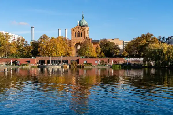 Herbstliche Bäume spiegeln sich im Wasser des Engelbeckens Berlin. Ein Café mit Außenbereich und eine Kirche mit Kuppel sind zu sehen.