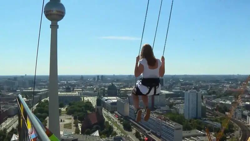 Frau schaukelt hoch über Alexanderplatz Berlin, mit Blick auf die Stadt und den Fernsehturm.
