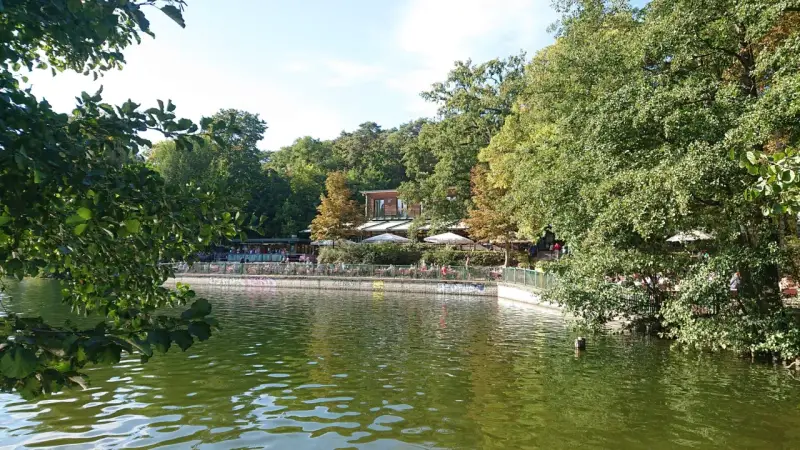 Grüne Bäume säumen den Schlachtensee Berlin, wo Menschen auf der Terrasse eines Restaurants sitzen.