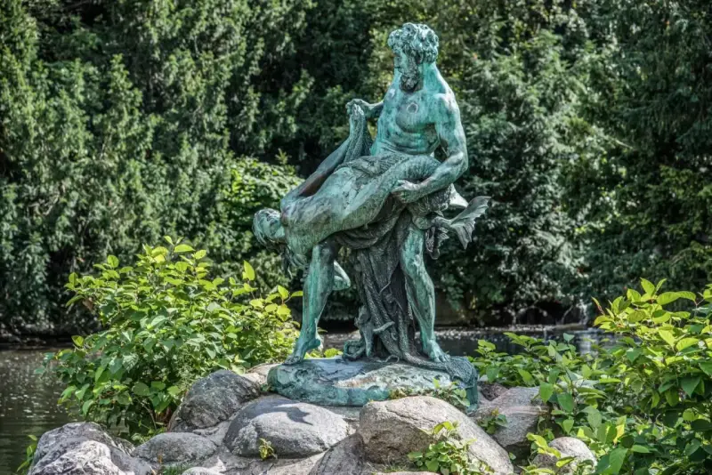 Bronzestatue eines Mannes, der eine Frau trägt, vor einem grünen Hintergrund mit einem kleinen Wasserfall in Berlin.