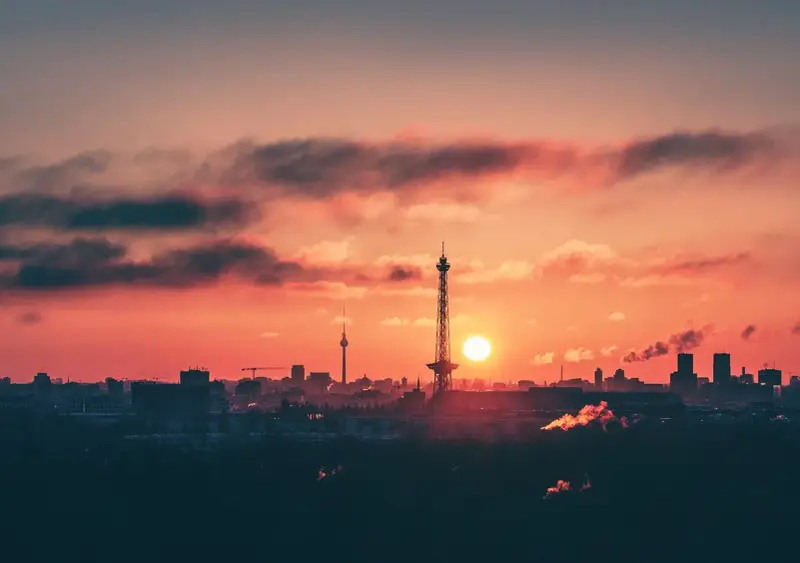 Berlin bei Nacht von oben: Die Sonne geht unter und taucht den Himmel in ein feuriges Rot. Silhouetten von Türmen und Gebäuden zeichnen sich ab.