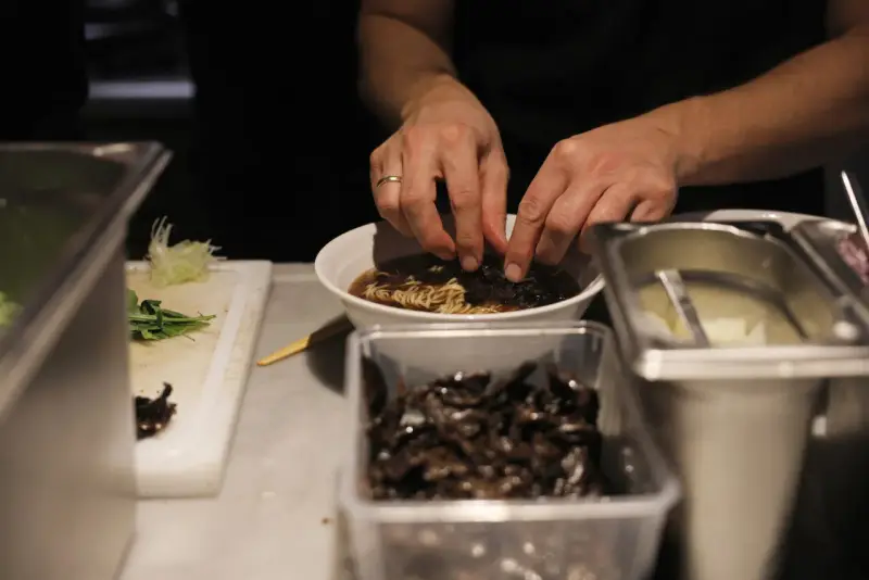 Hände garnieren eine Schüssel mit Ramen-Nudeln in Berlin. Daneben liegen Zutaten bereit.