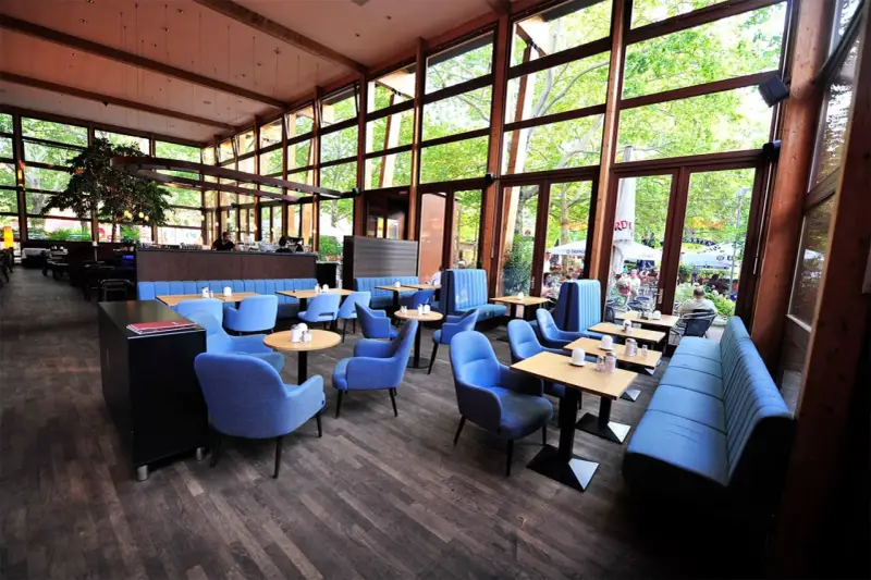 Gemütliches **Cafe Wilmersdorf** mit blauen Sesseln und Bänken, Holzboden und großen Fenstern, die auf eine grüne Terrasse blicken.