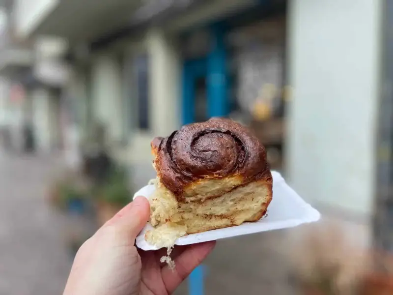 Hand hält ein saftiges Zimtschnecken-Stück vor einer unscharfen Berliner Kulisse.